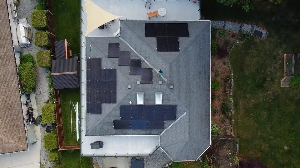 Top down drone view of black solar panels on a hipped shingle roof of a Lower Mainland home, with an installer on the roof
